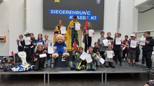 Siegerehrung der Klasse K1 bei der Baden-Württembergischen Kartslalom-Meisterschaft 2026 in Backnang: Nachwuchsfahrer mit Urkunden und Pokalen auf dem Podium, der Sieger mit Meisterumhang auf Platz 1, links ein blau-weißes Jugendkart