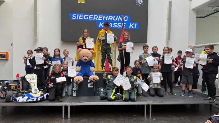 Siegerehrung der Klasse K1 bei der Baden-Württembergischen Kartslalom-Meisterschaft 2026 in Backnang: Nachwuchsfahrer mit Urkunden und Pokalen auf dem Podium, der Sieger mit Meisterumhang auf Platz 1, links ein blau-weißes Jugendkart