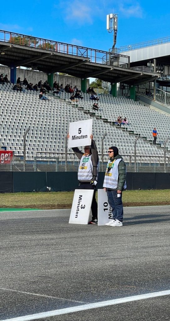 Helfer mit den Tafeln „5 Minuten“ und „10“ in der Startaufstellung vor der Tribüne